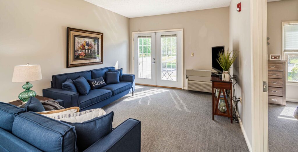 Interior image of a resident's living room with comfortable furniture and colorful artwork. There is a double door leading out to a patio space on the far wall.