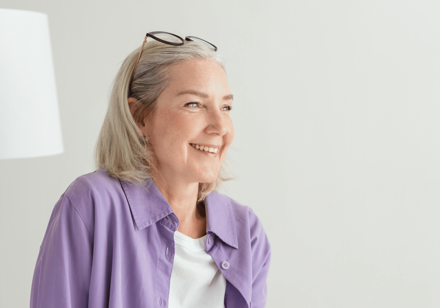 Smiling older woman with gray hair in a lavender shirt, glasses resting on her head, indoors by a soft light.