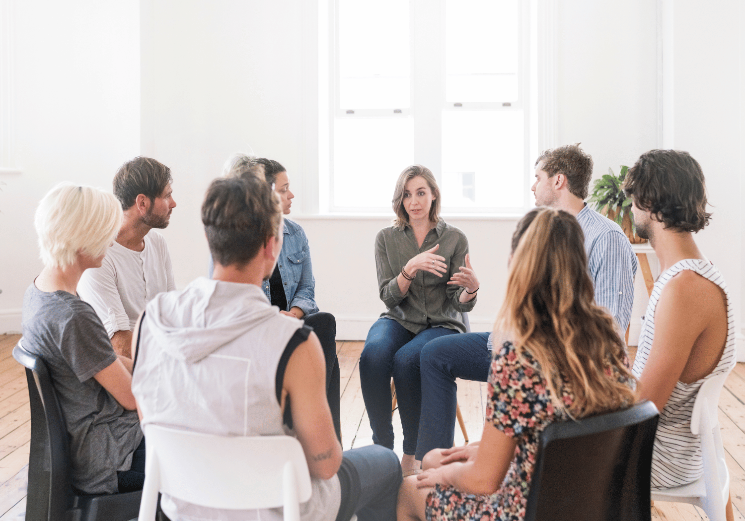A woman speaks while seated in a circle with eight others in a bright room, suggesting a group discussion or workshop.