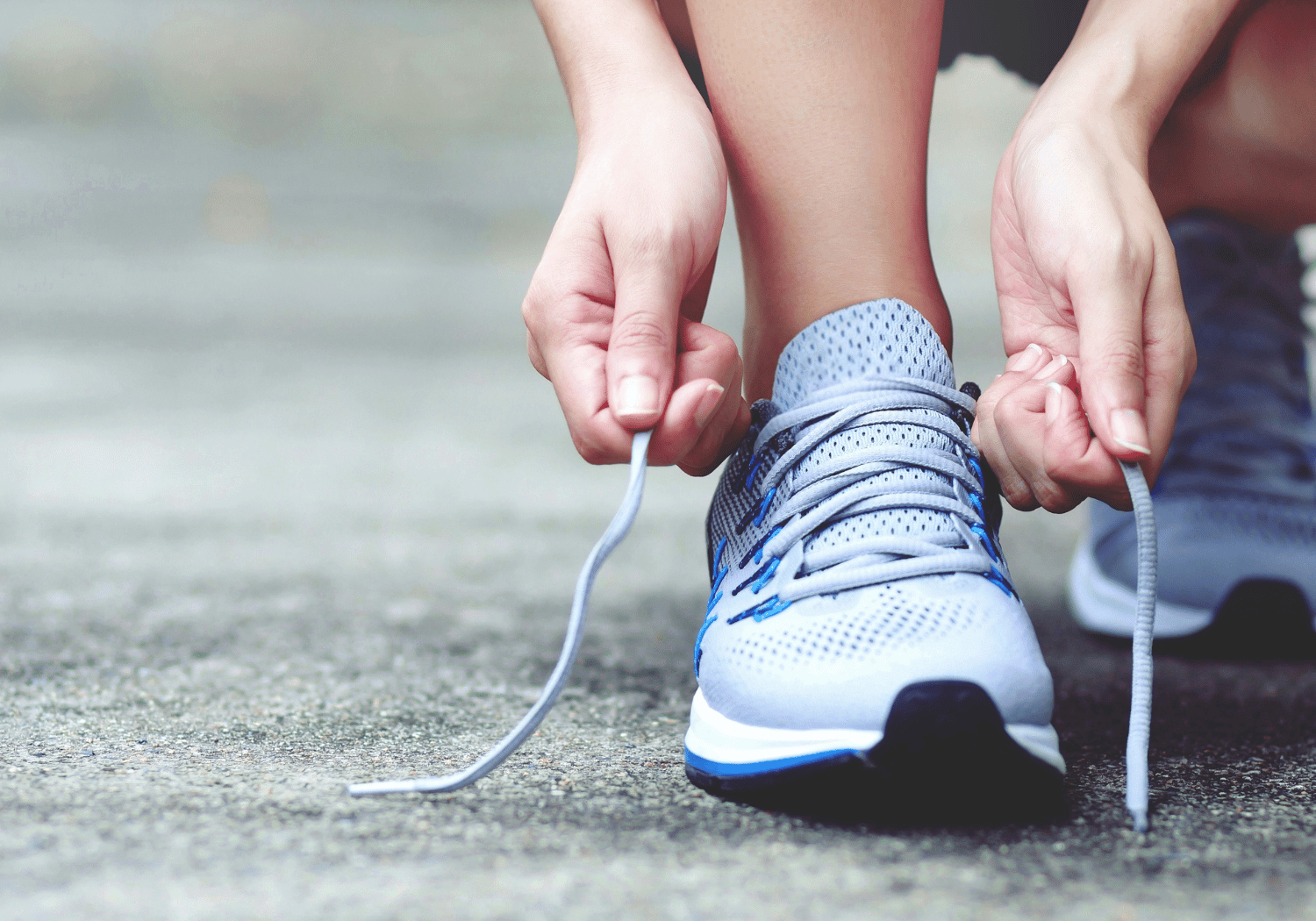 Close-up of hands tying the laces on a light blue running shoe on a pavement surface.